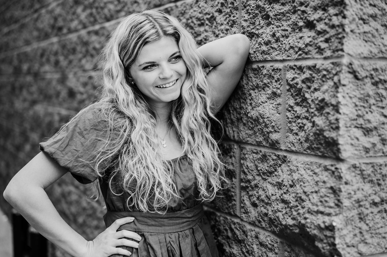 Black and white portrait of a person in casual dress leaning against a textured stone wall while smiling at camera.