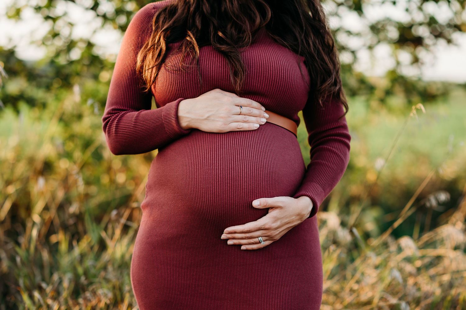A pregnant woman in a burgundy dress cradles her baby bump while standing in a sunlit outdoor setting at Prophetstown State Park in Indiana.