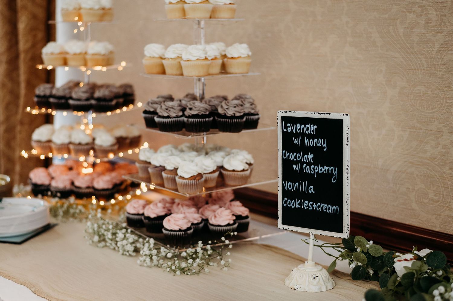 Multi-tiered display of cupcakes with baby's breath flowers and chalkboard sign at wedding dessert table.