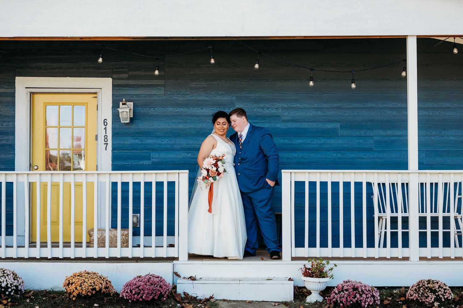 Blue-sided building with white porch railing and yellow door decorated with colorful fall flowers.