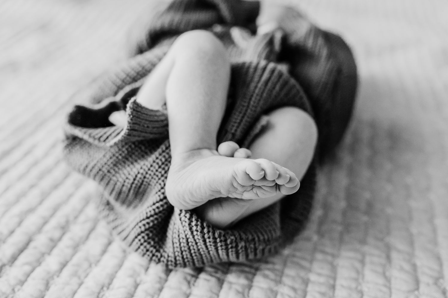 A black and white image showing small newborn bare feet peeking out from a cozy knit sweater in Indiana home.