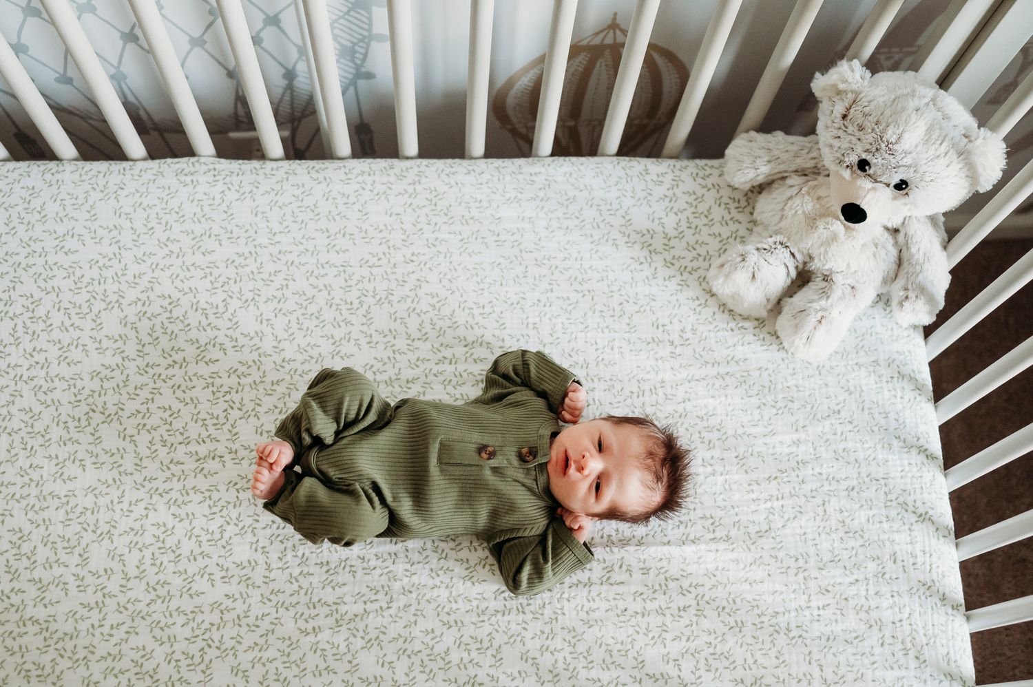 A baby in a green onesie lies in a crib with a plush teddy bear on a light gray sheet.
