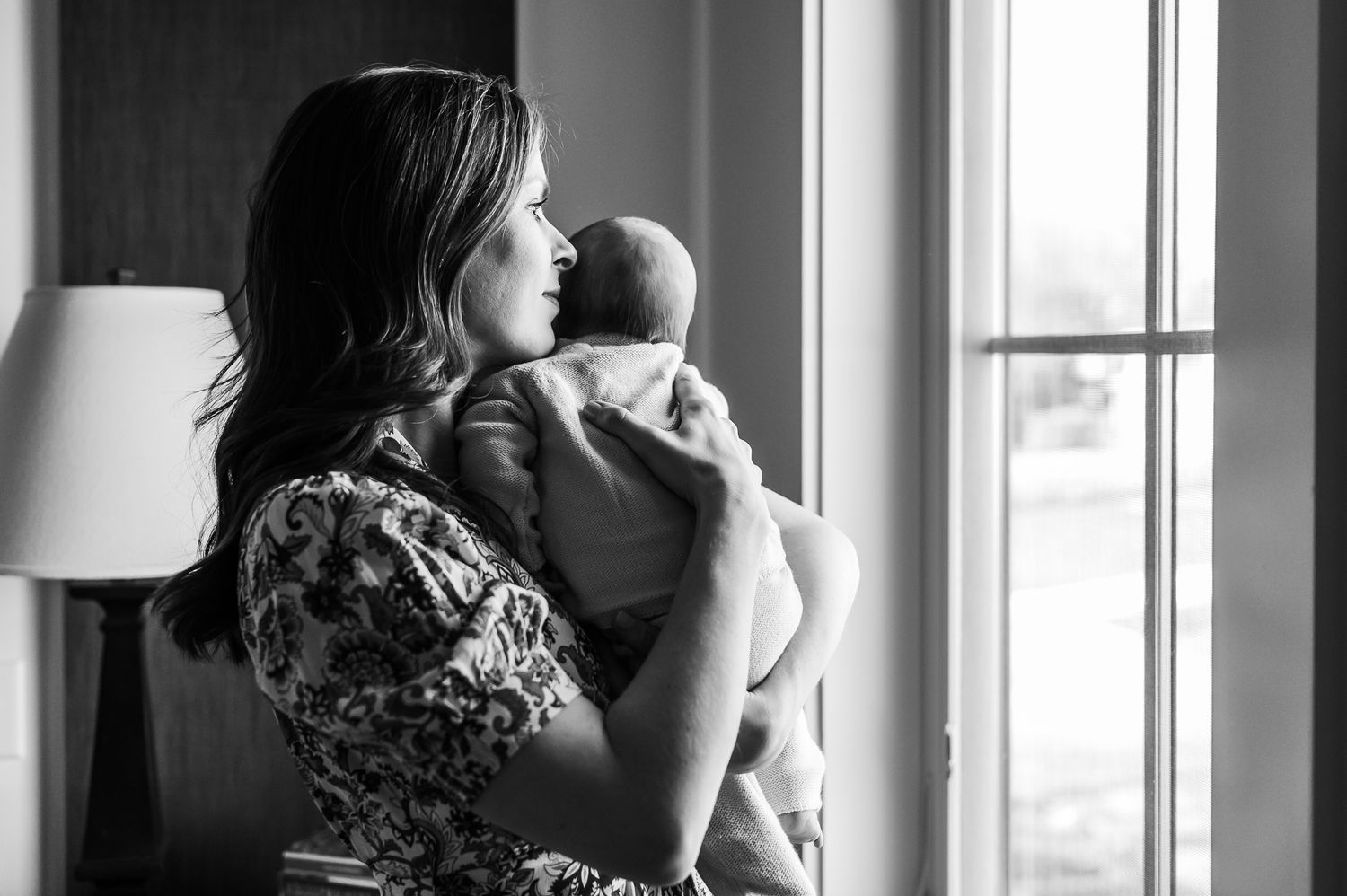 A woman in a floral dress holds a newborn baby while standing by a bright window in a black and white photograph.