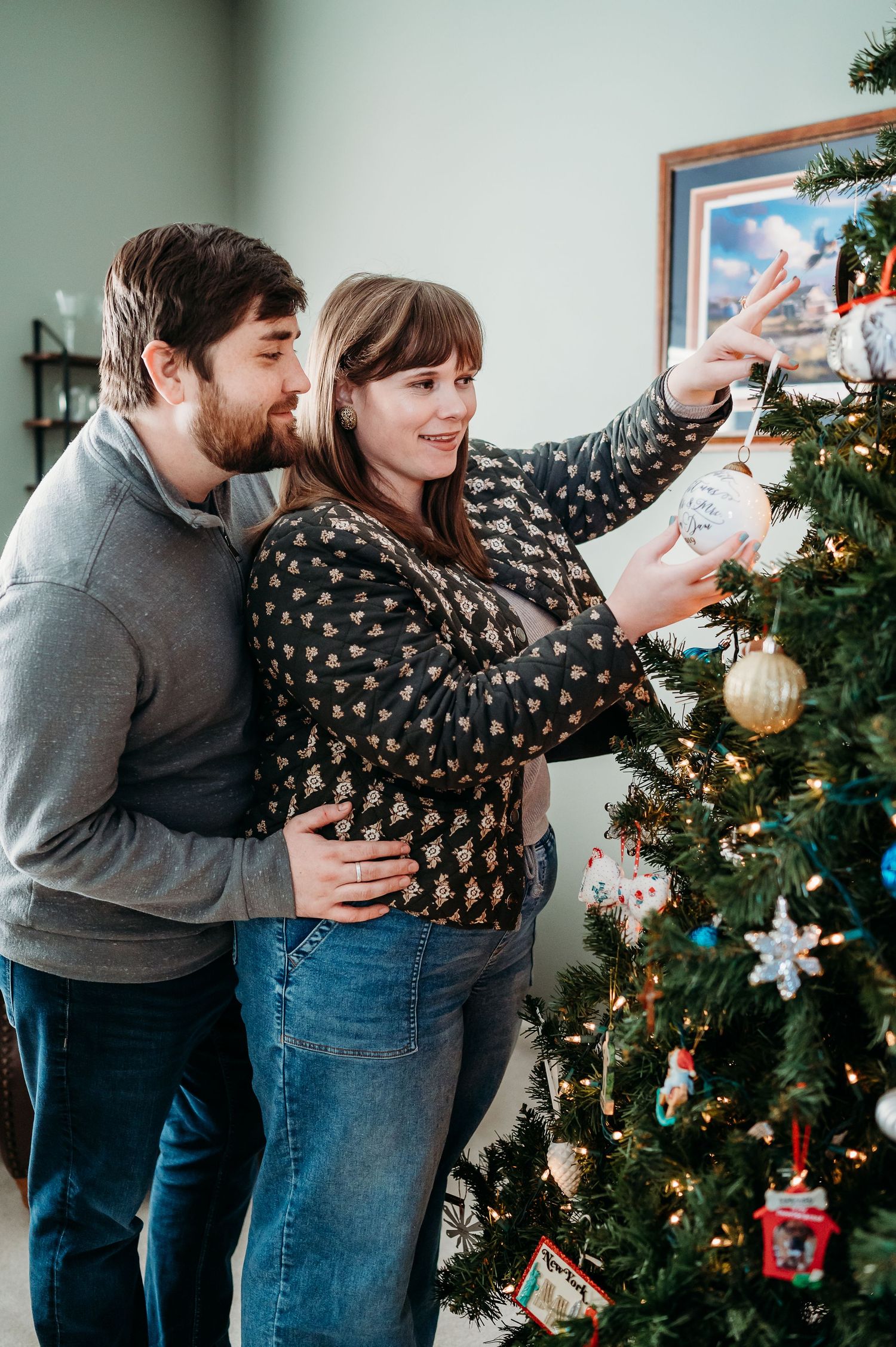 Couple decorating a Christmas tree together, hanging ornaments on a lit green tree indoors.