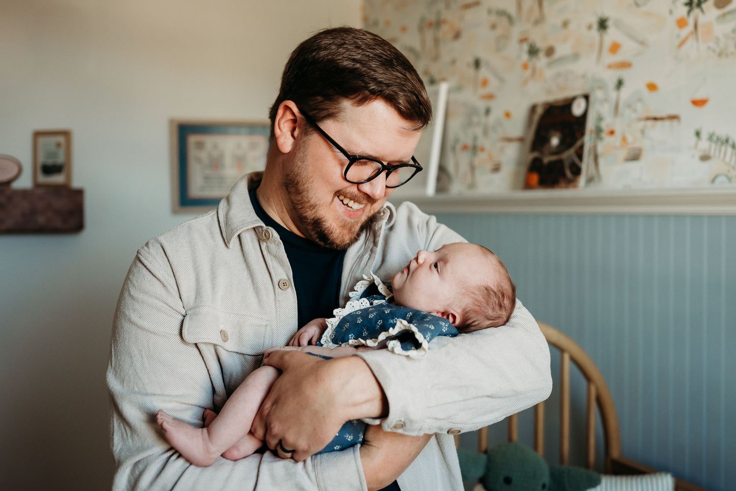 A father with glasses tenderly holds his newborn baby in a cozy nursery room.