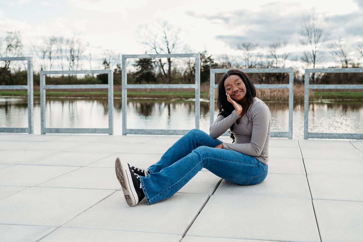 Girl sits casually on dock near a serene lake with  railings on a cloudy day during senior photoshoot.