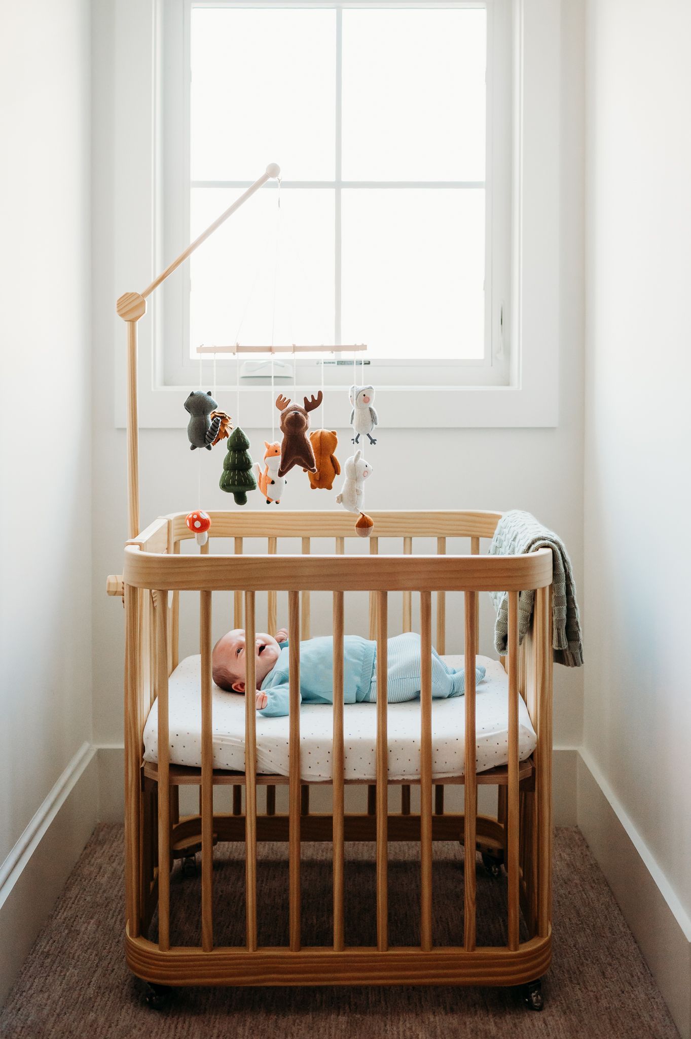 Baby boy sleeps in wooden oval baby crib with mobile featuring plush toys near a bright window in a minimalist nursery setting.
