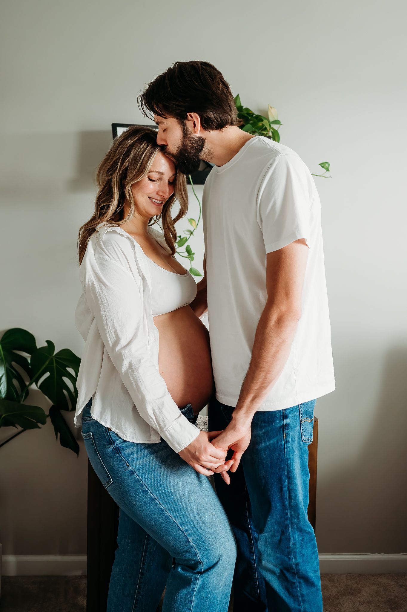 A tender maternity moment captured indoors with a romantic embrace near houseplants against a light gray wall.
