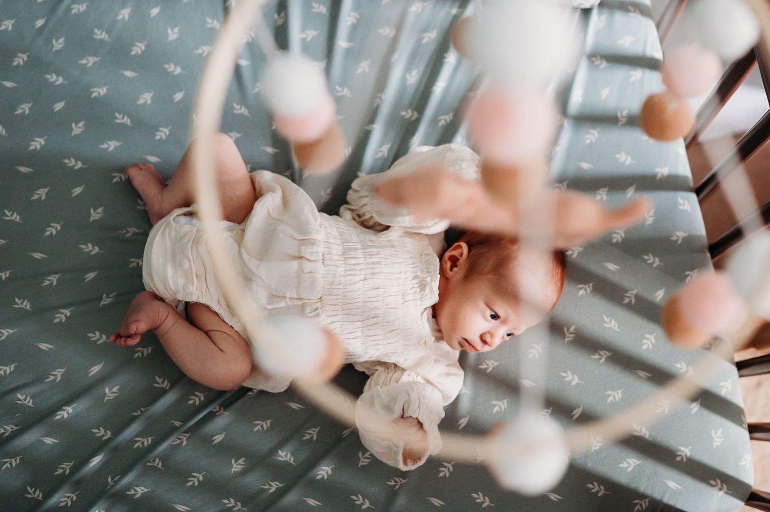 Baby wearing white lace dress lies on textured grey blanket while reaching up with tiny hands.