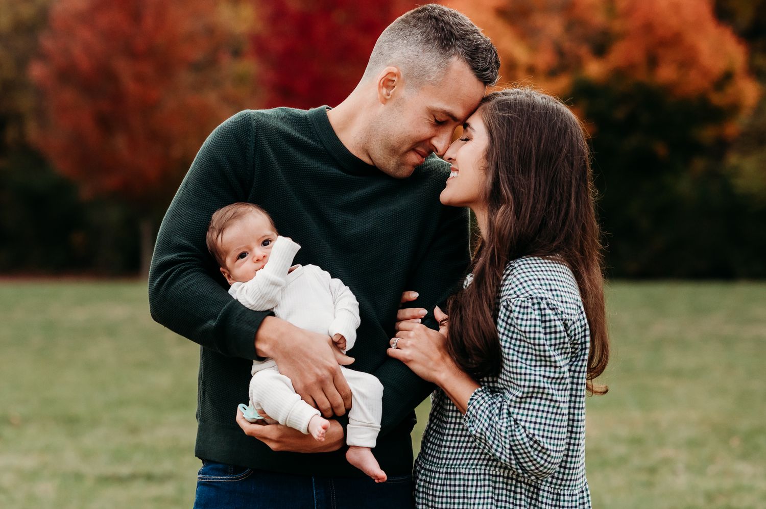 An Avon, Indiana, couple holds a newborn baby outdoors during autumn with warm orange foliage in the background.