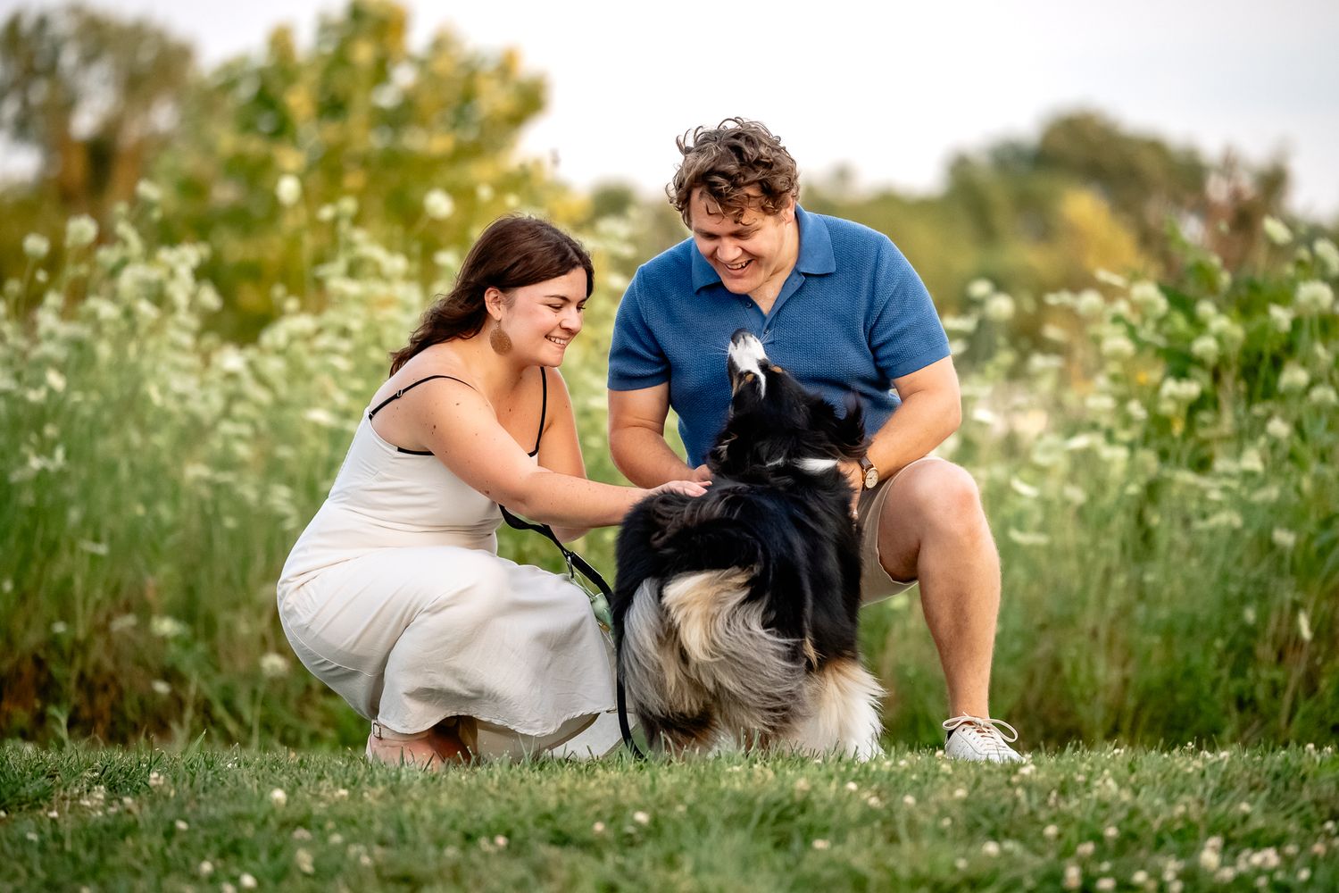 A couple smiles while petting Australian Shepherd dog outdoors in a lush green meadow with wildflowers during photoshoot with Sara House.