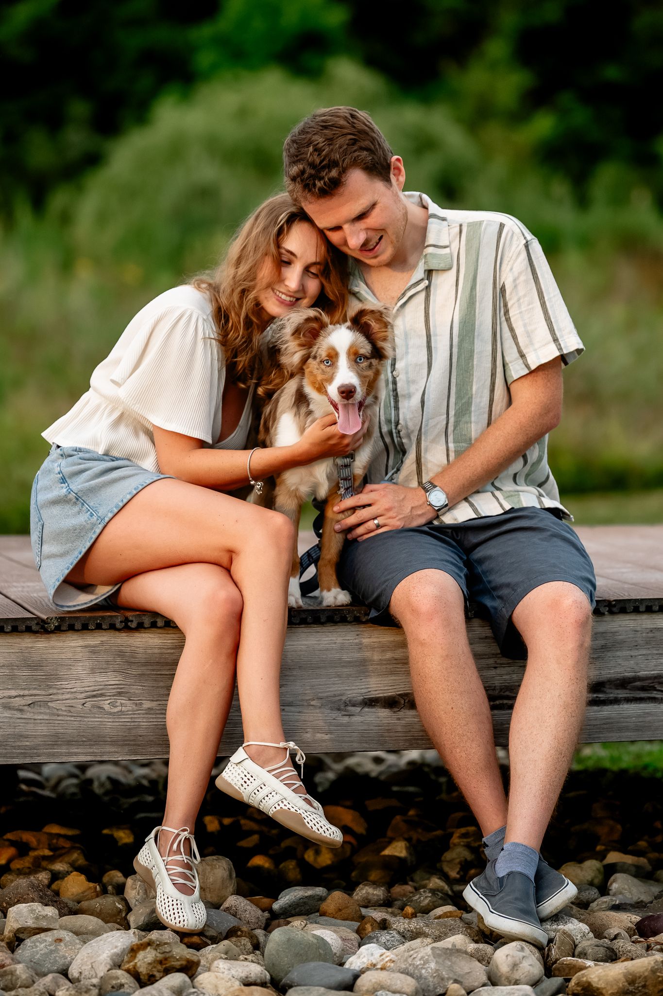 Couple sitting on wooden dock with their Australian Shepherd dog outdoors during Plainfield, Indiana dog photoshoot.