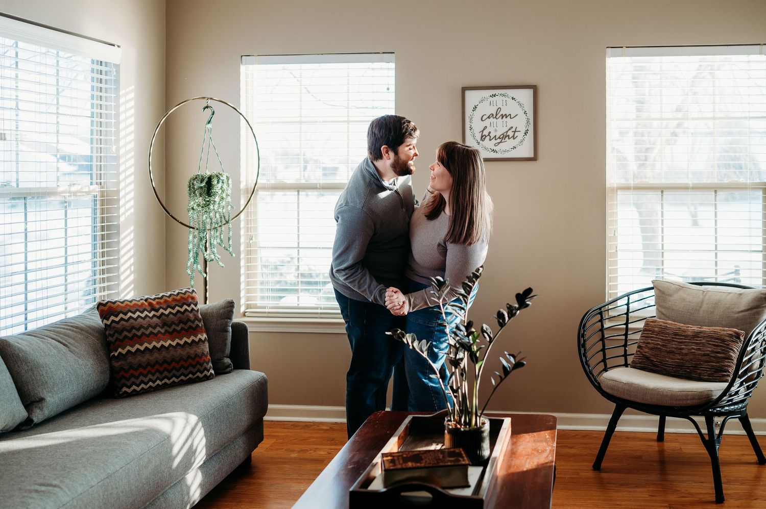 A couple shares a romantic embrace while standing in a sunlit living room with hardwood floors during an Indianapolis in-home photo session.