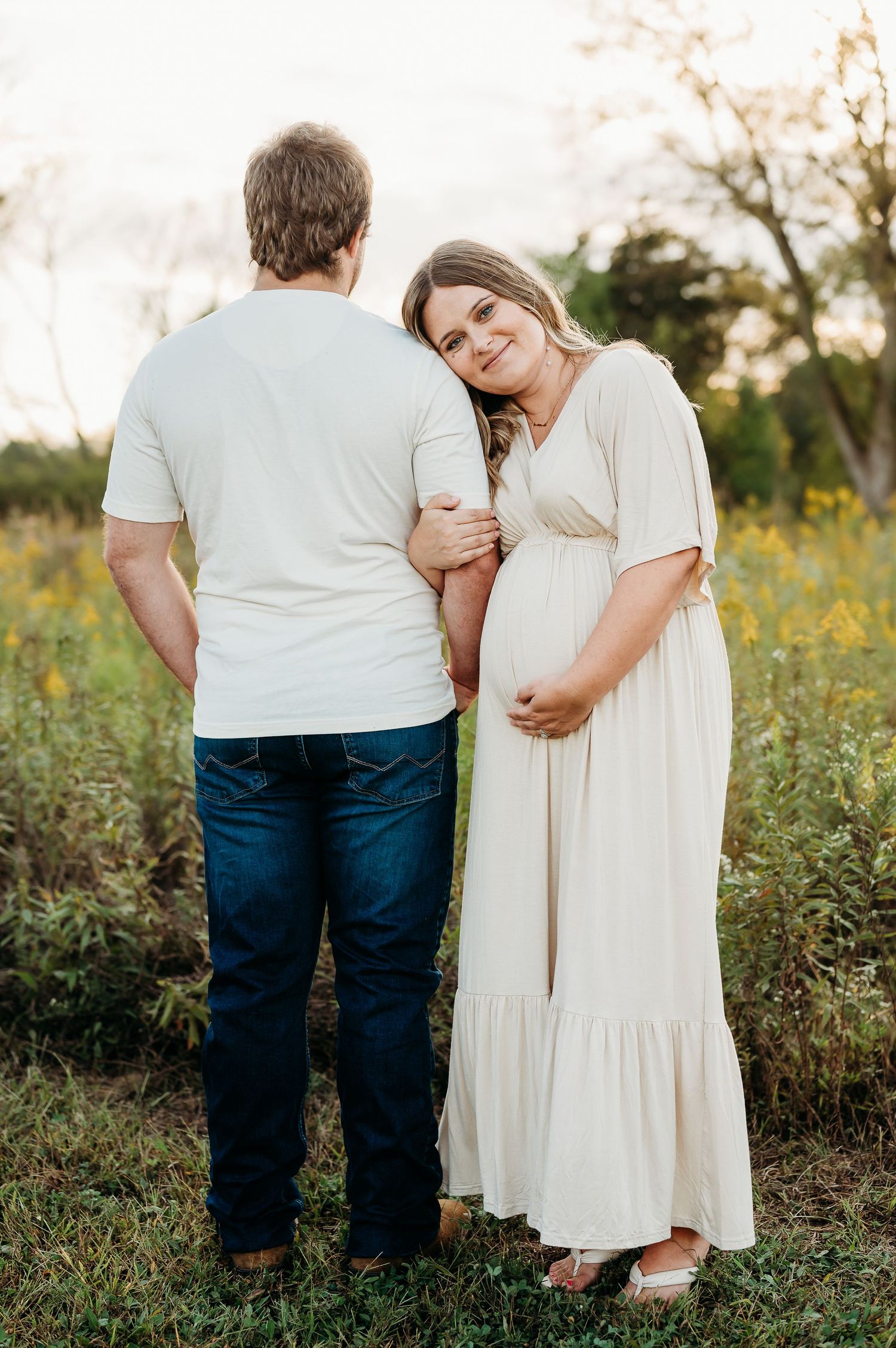 An expecting Indianapolis mother in cream dress embraces husband in denim during sunset maternity photoshoot.