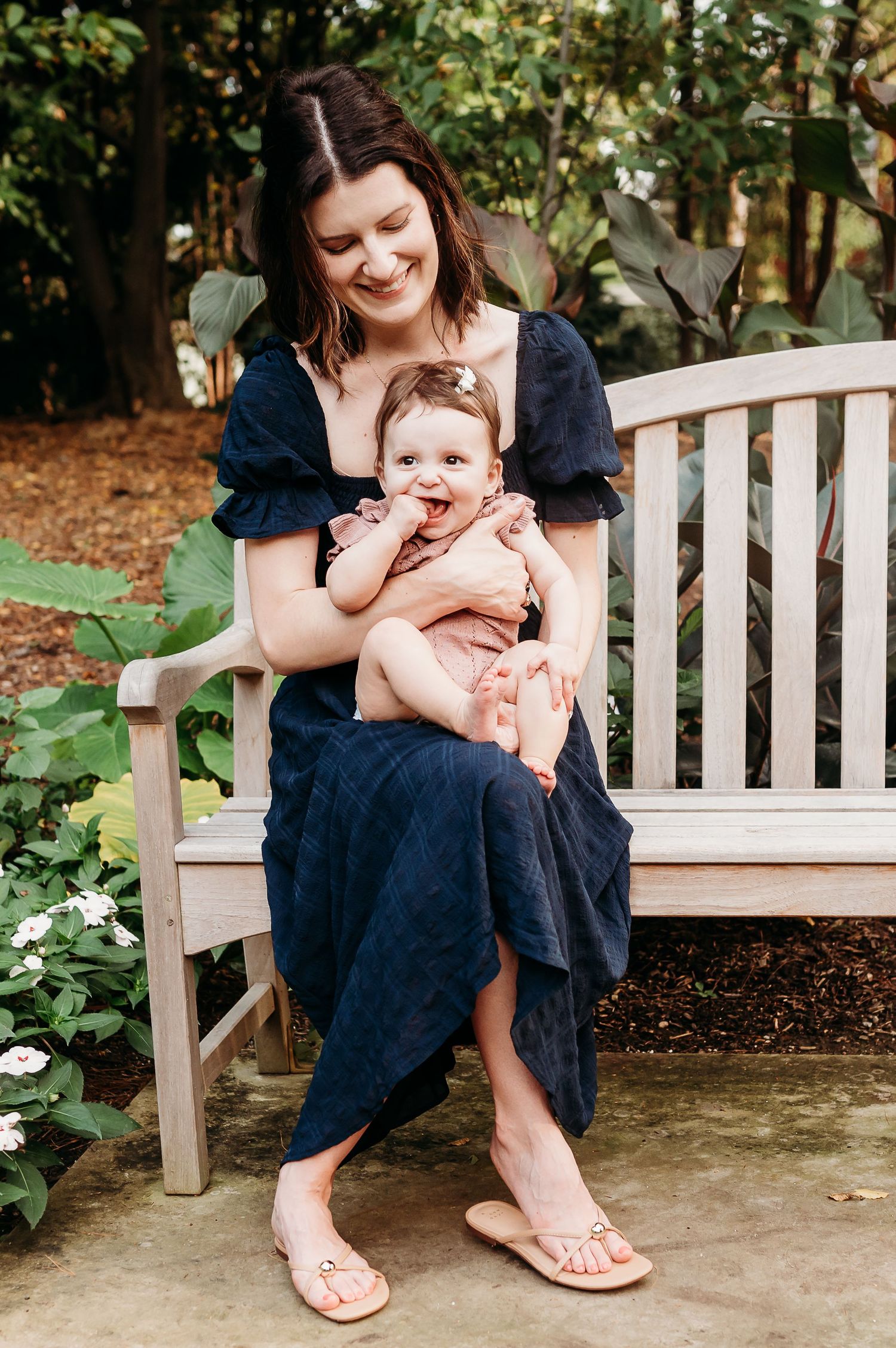 A heartwarming outdoor portrait of a mom and baby girl on a wooden bench surrounded by lush garden greenery at Newfields in Indianapolis.