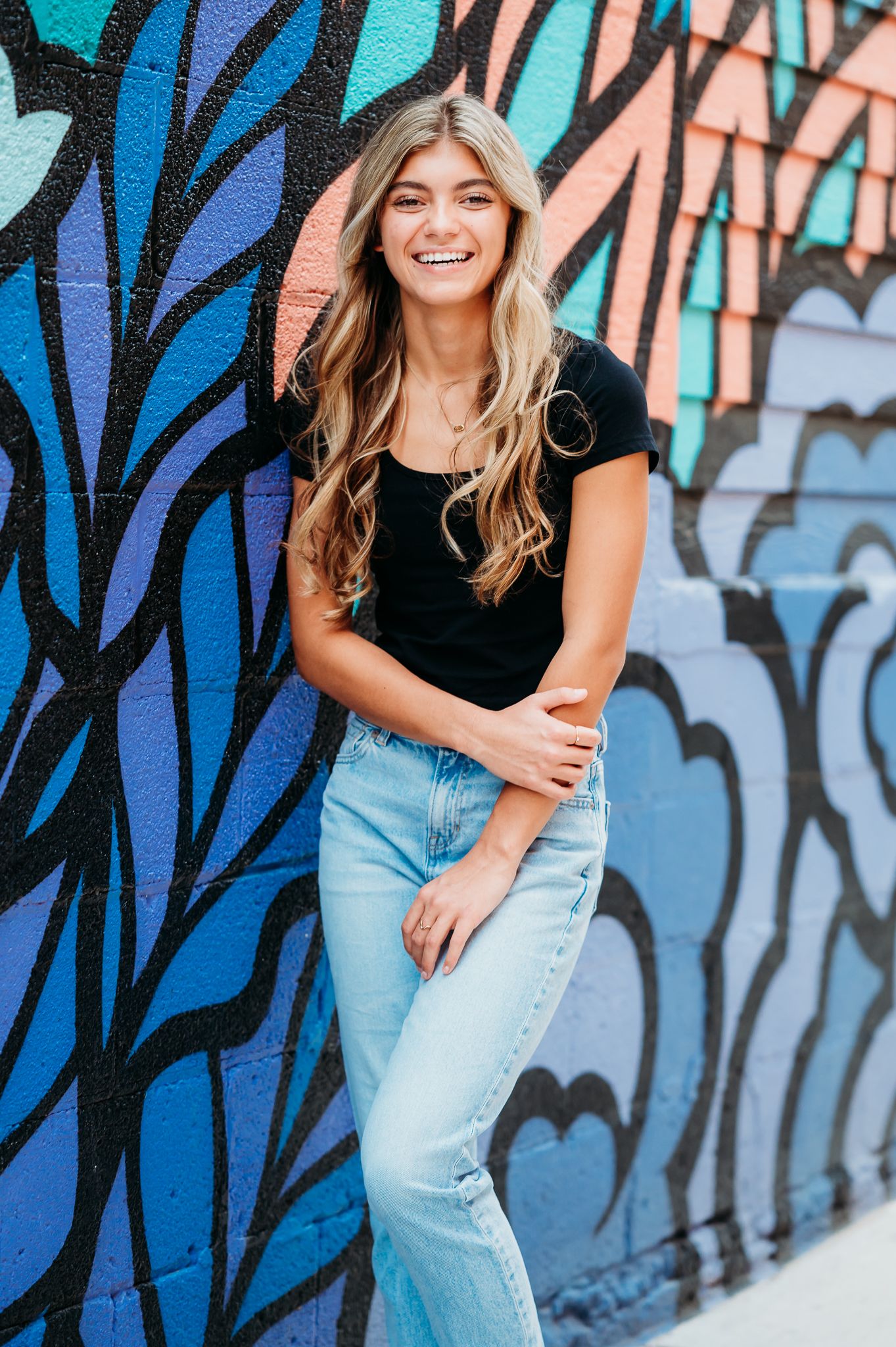 A person in a black top and light jeans poses cheerfully against a colorful graffiti wall with blue and pink designs.