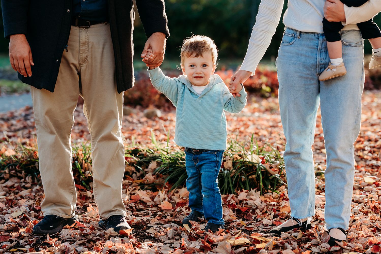 A toddler in a blue shirt and jeans walks while holding hands with parents on a path covered in autumn leaves in Plainfield, Indiana.