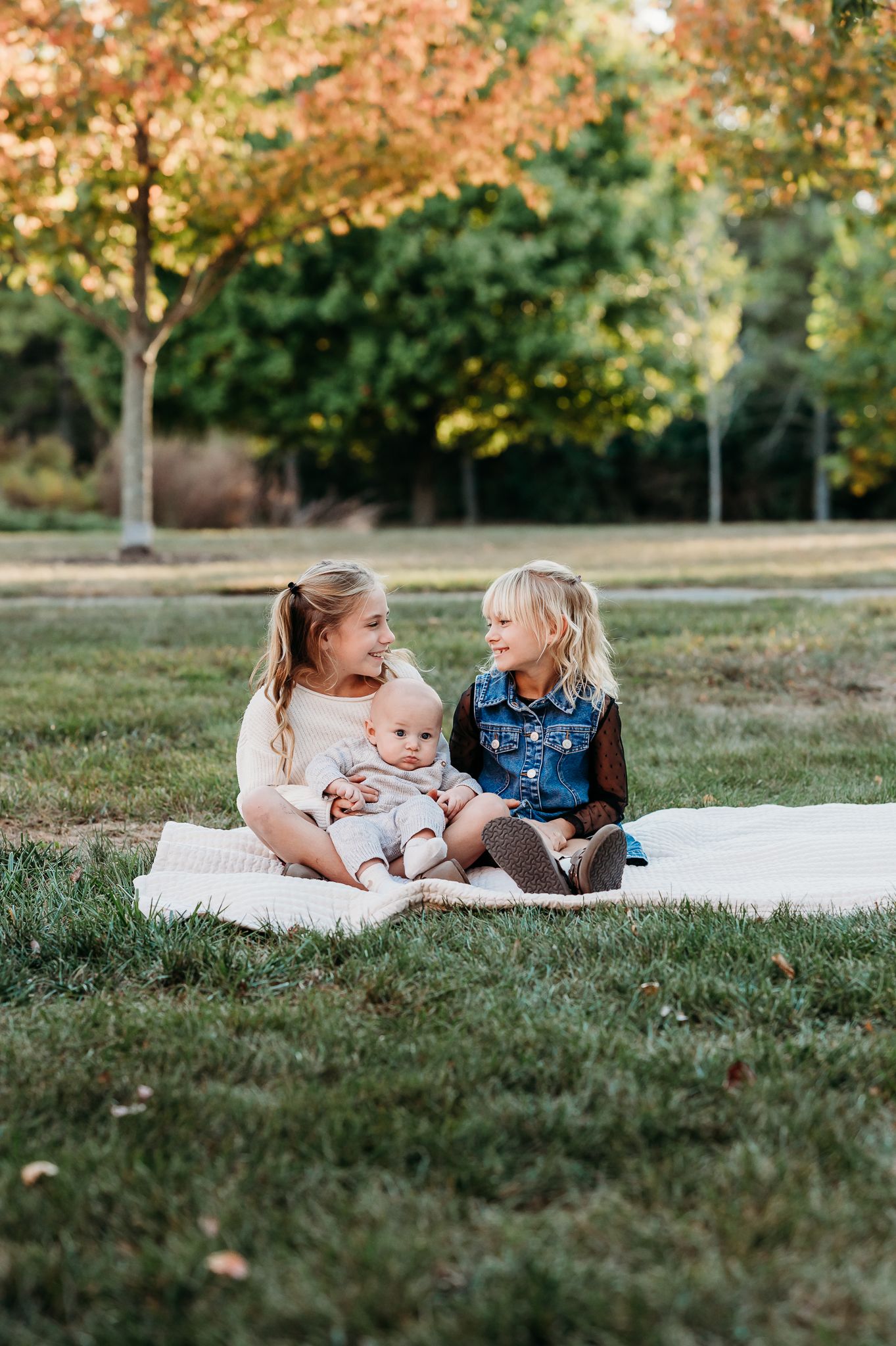 Sisters together with baby brother on blanket in during fall family photoshoot in Plainfield, Indiana, with golden trees in the background.