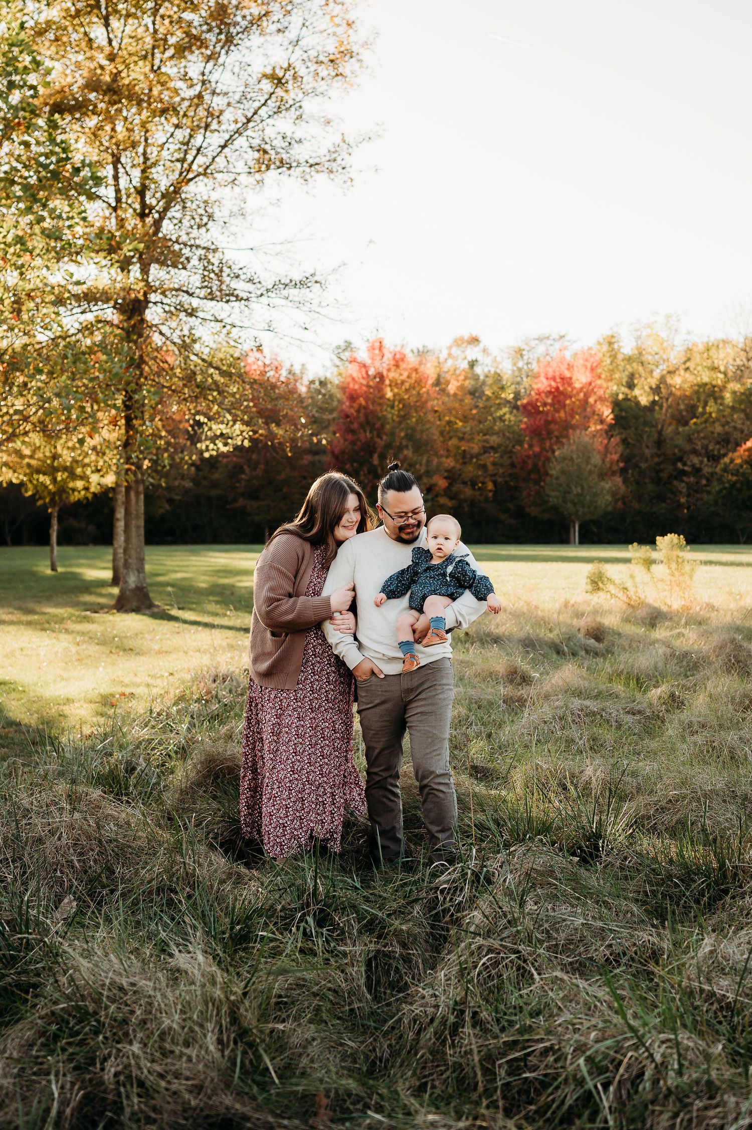 A family stands together in a grassy field during golden hour with autumn trees glowing in the background.