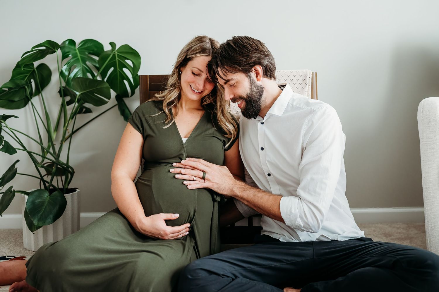 Pregnant woman in green dress with bearded partner tenderly touching baby bump during maternity photo session indoors.