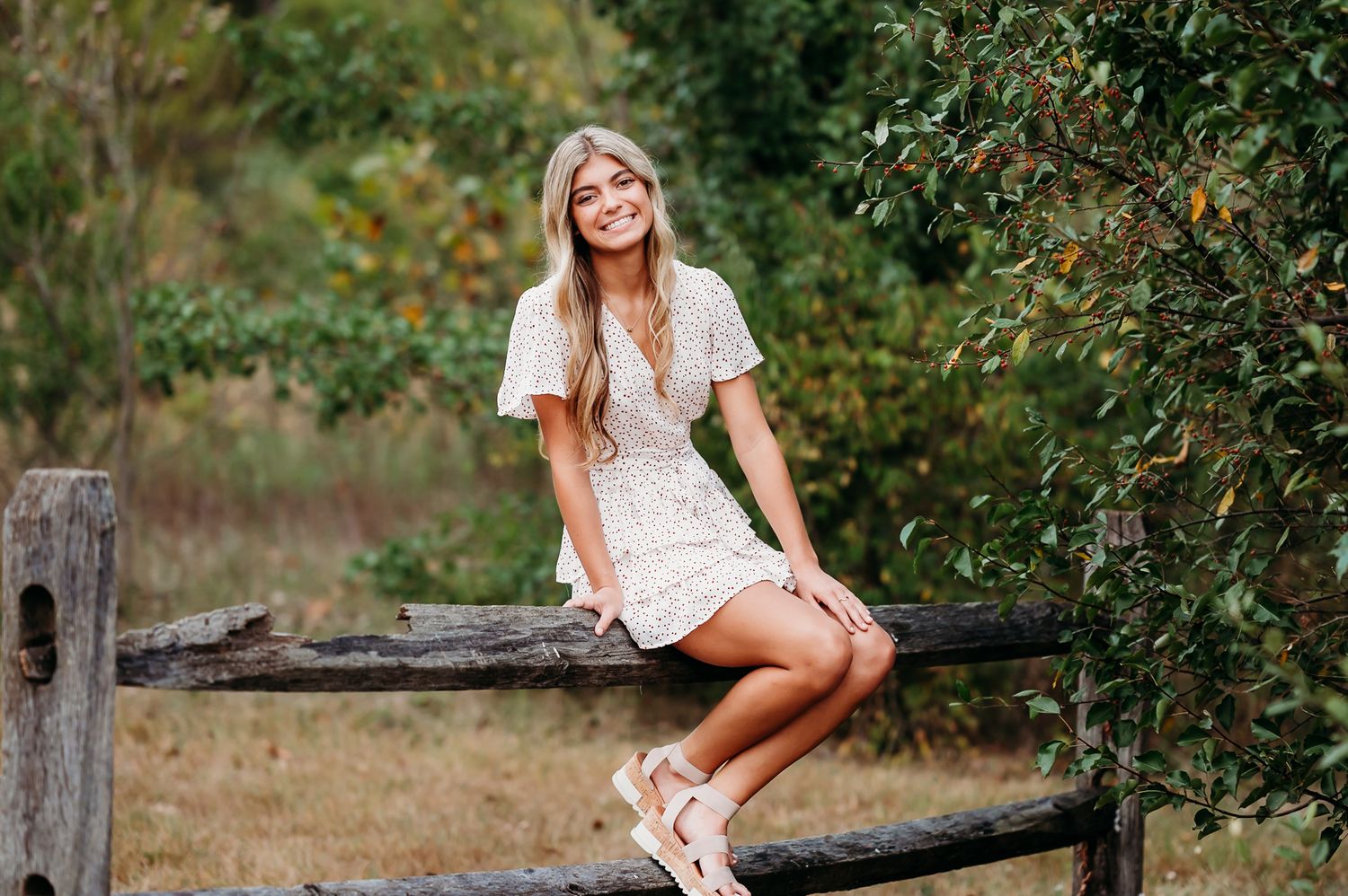 Woman in white lace dress sits casually on rustic wooden fence surrounded by green foliage in outdoor setting.