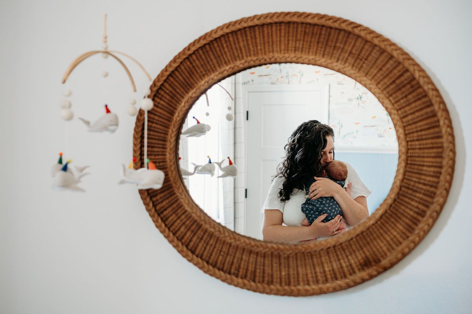 Mother cradling newborn baby reflected in a round wicker mirror in a nursery with a decorative mobile.