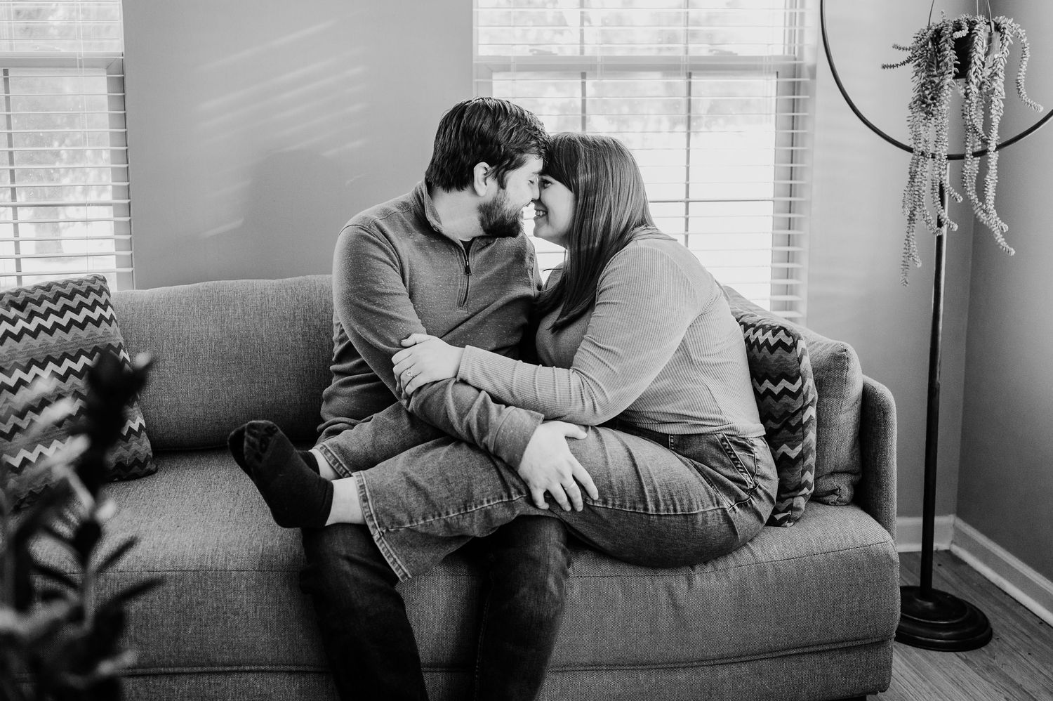 Black and white photo of a couple sharing a tender kiss while sitting together on a cozy gray sofa in a living room.