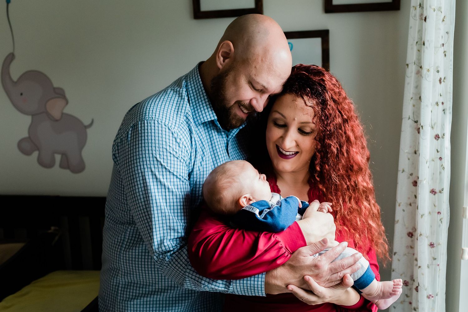 A couple embraces tenderly while holding their newborn baby against a white wall with elephant decor.