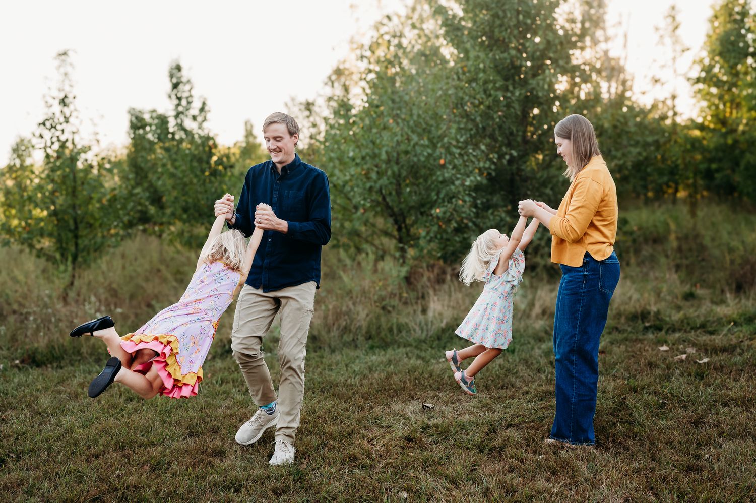 Two adults swing children playfully in a grassy field during golden hour while enjoying outdoor family time together in Avon, Indiana.
