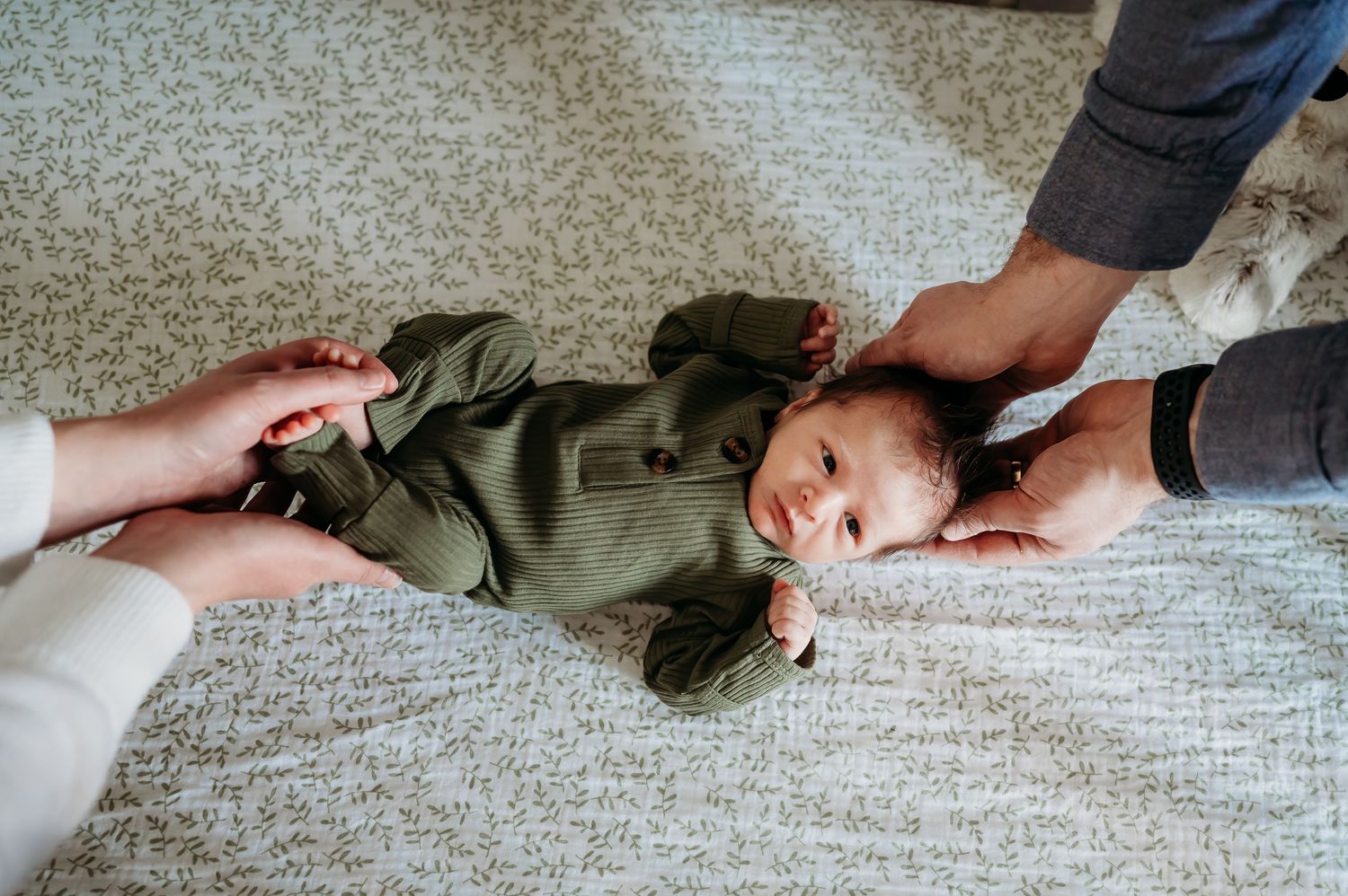 A baby wearing a green outfit lies on a gray carpet while hands reach to tickle them.