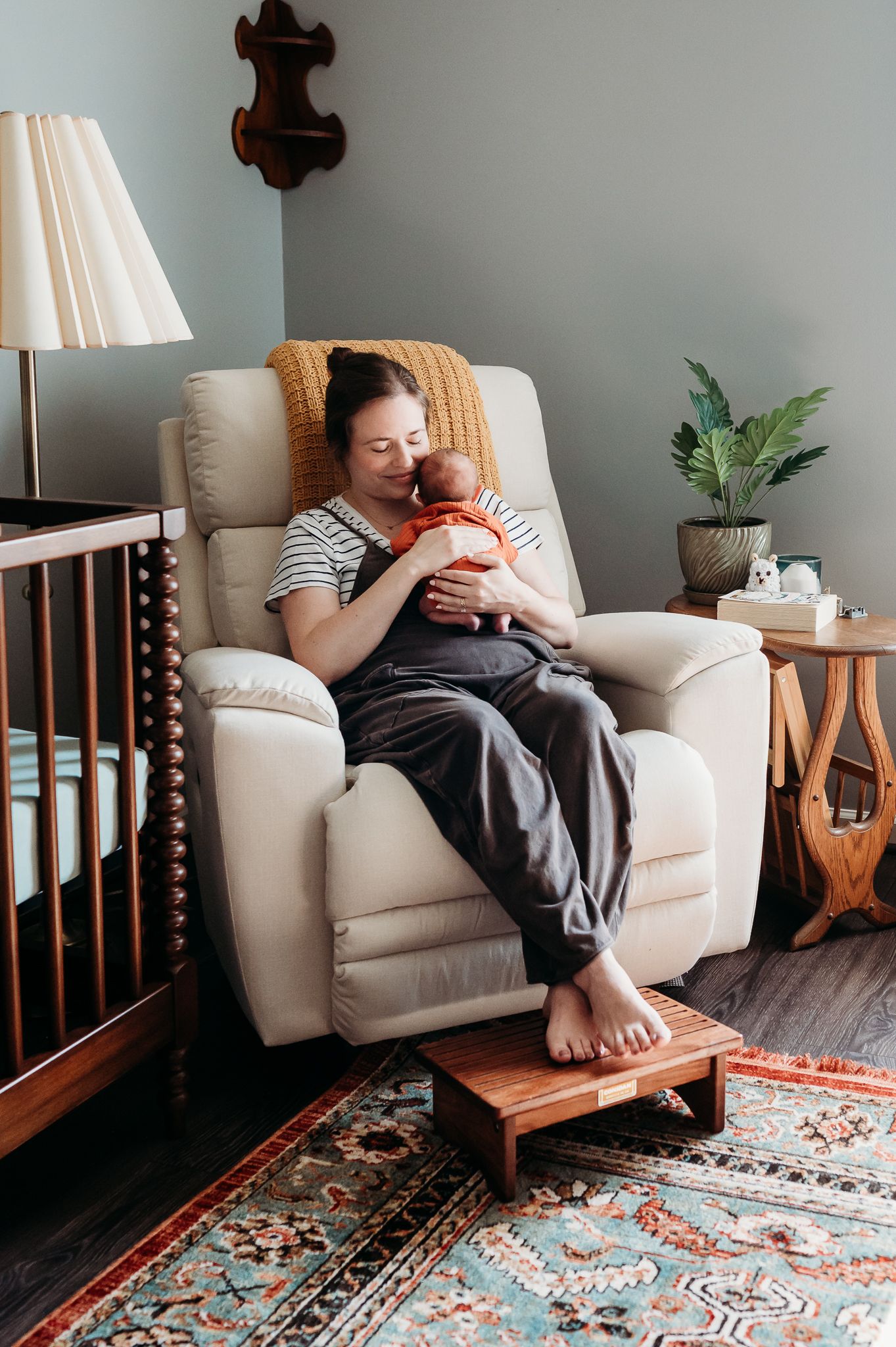 A cozy Indiana nursery features a recliner, wooden crib, potted plant and patterned area rug in warm earth tones with mom rocking baby.