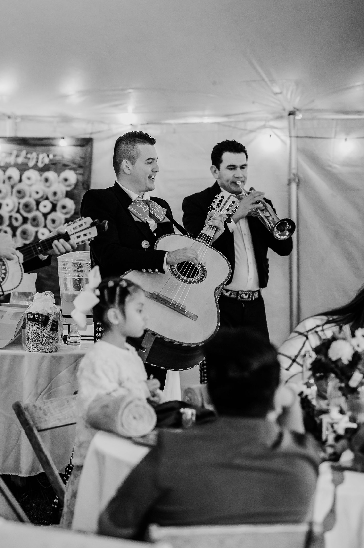 A mariachi band performs at an indoor event with musicians playing guitars and trumpets in black and white photography.