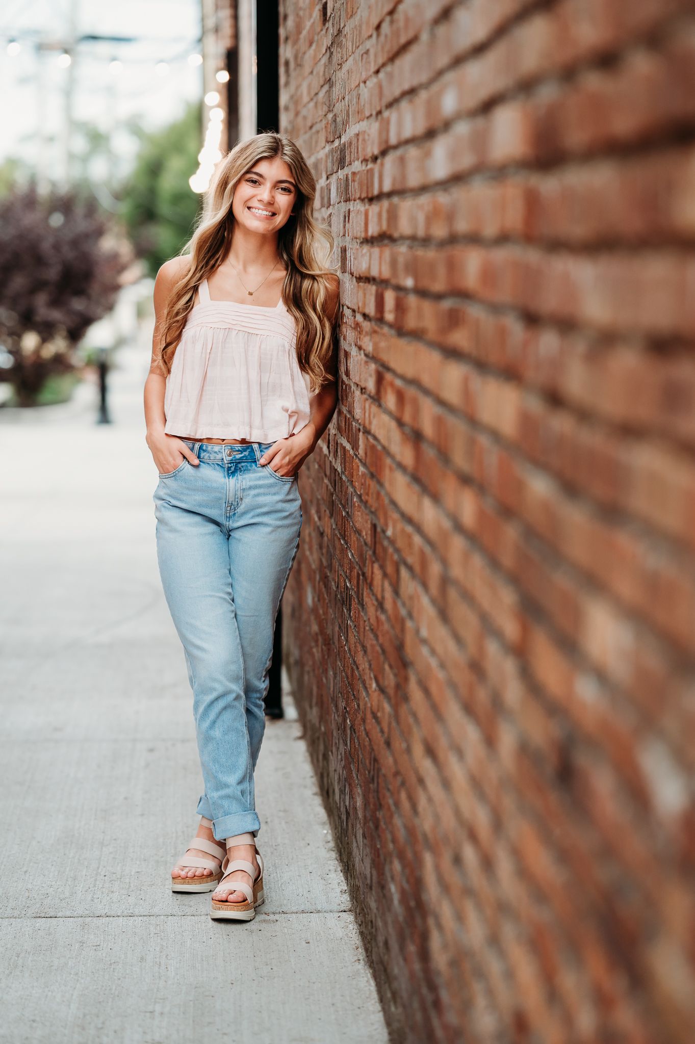 Young woman in a white summer top and light blue jeans poses casually against a brick wall on a city sidewalk.