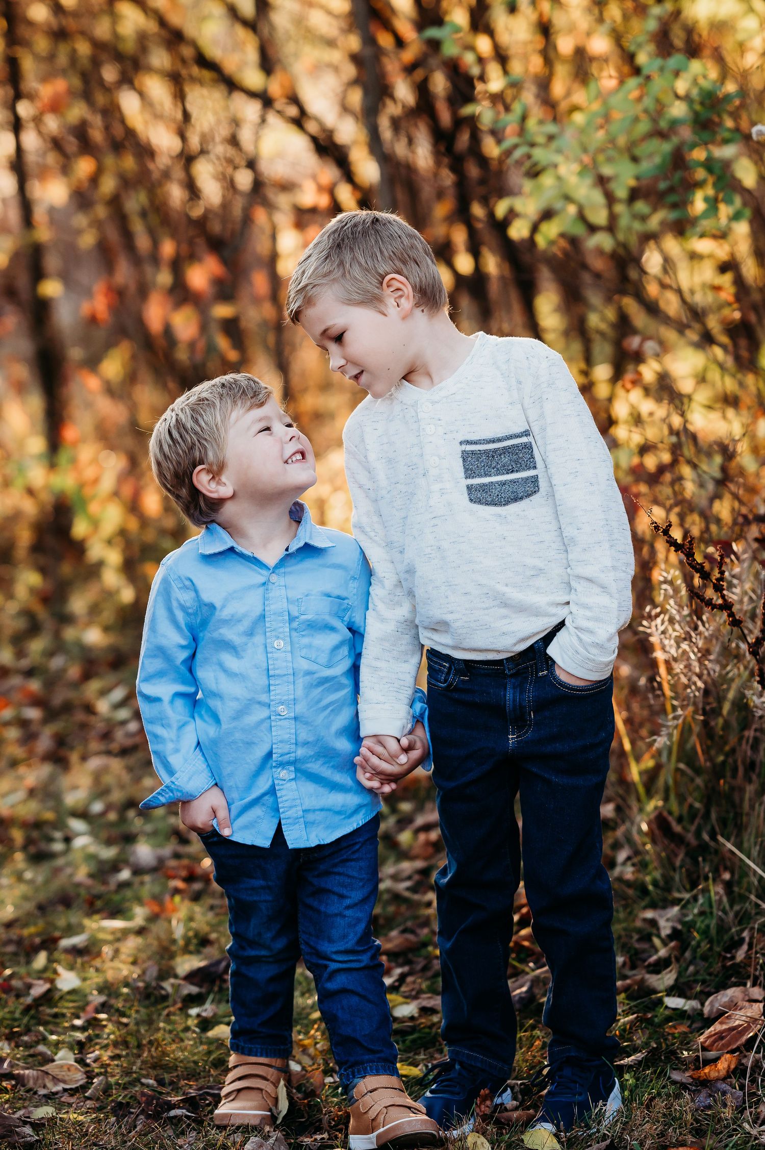 Two brothers in blue and white outfits share a playful moment together in a sunlit autumn forest setting in Fishers, Indiana.