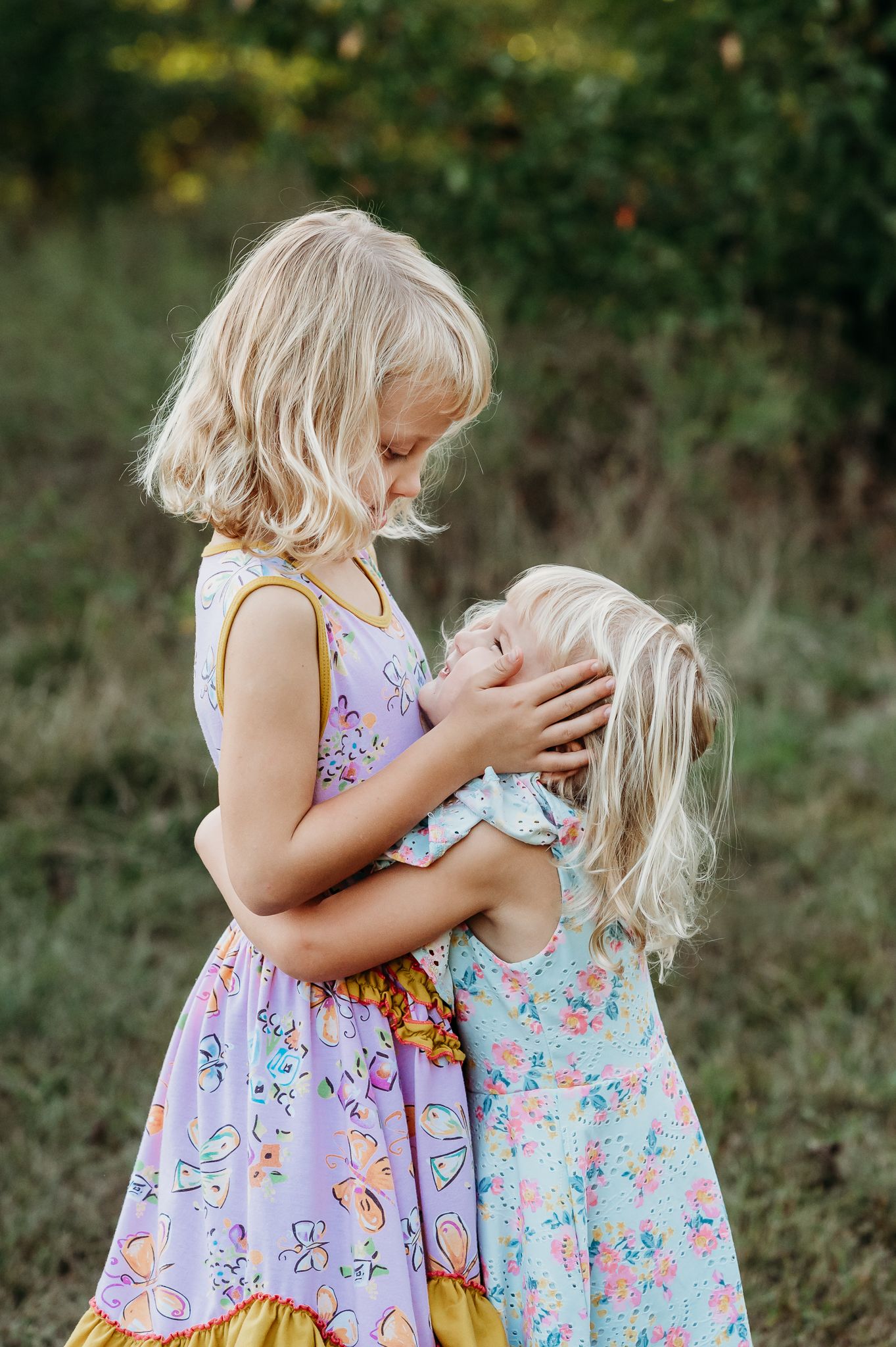 Two young children in floral summer dresses share a sweet sisterly moment outdoors in a field at Avon's Washington Township Park.