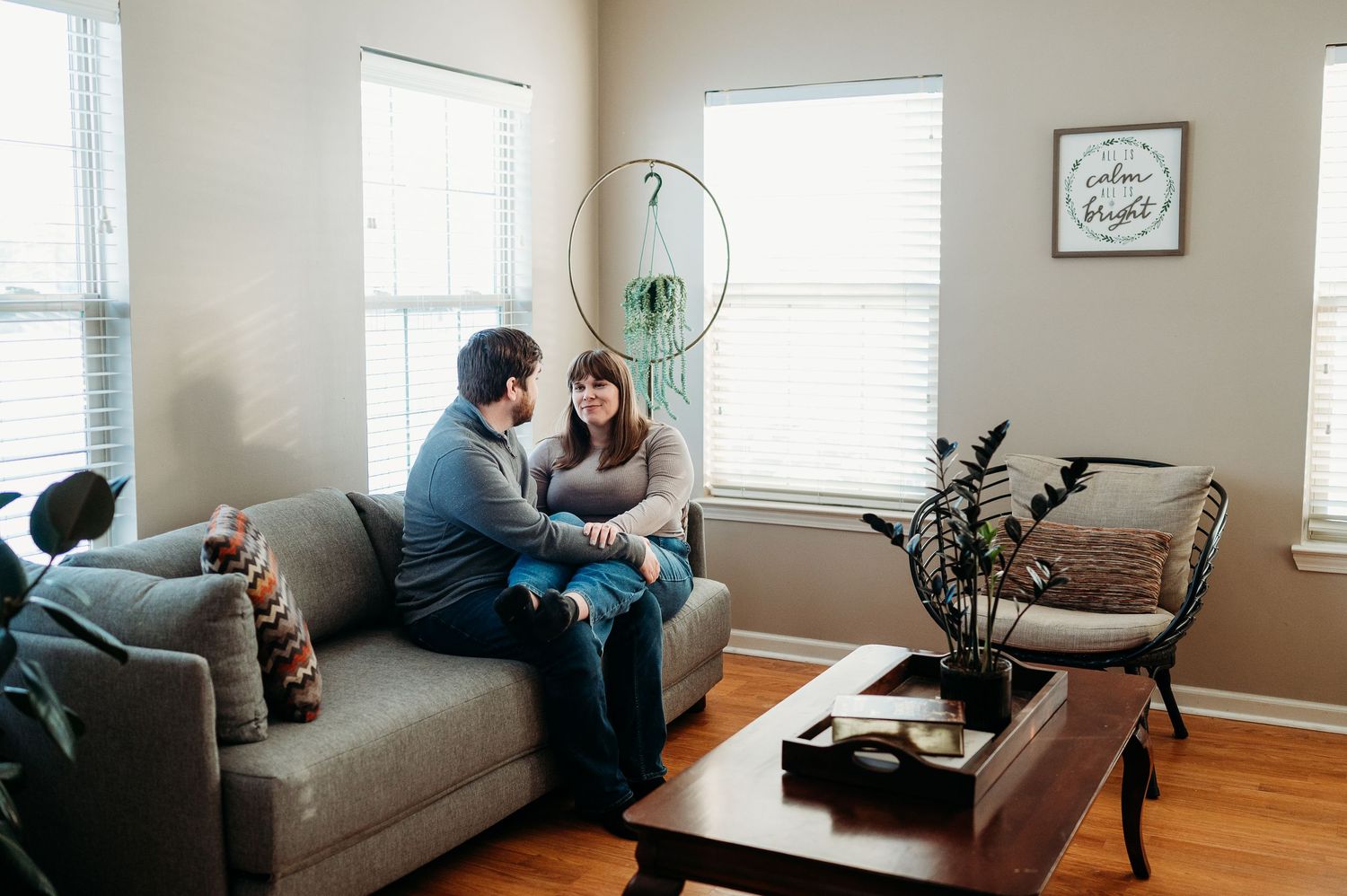 A couple sits together on a gray sofa in a bright, cozy living room, smiling and holding hands.