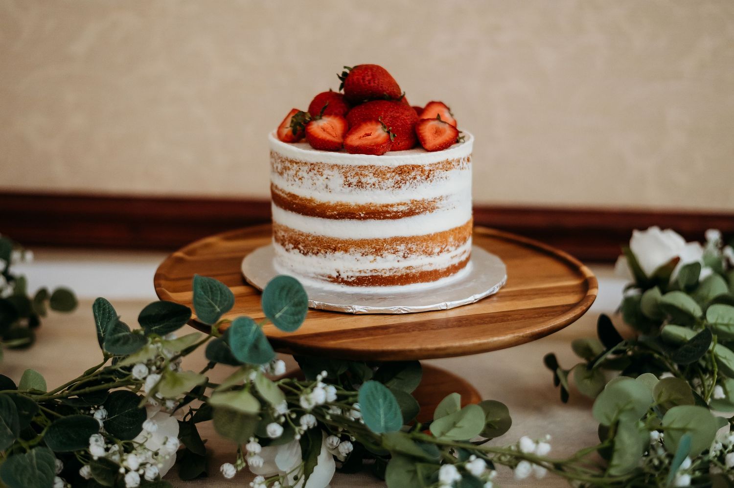 Minimalist wedding cake decorated with fresh strawberries and eucalyptus leaves on a wooden stand.