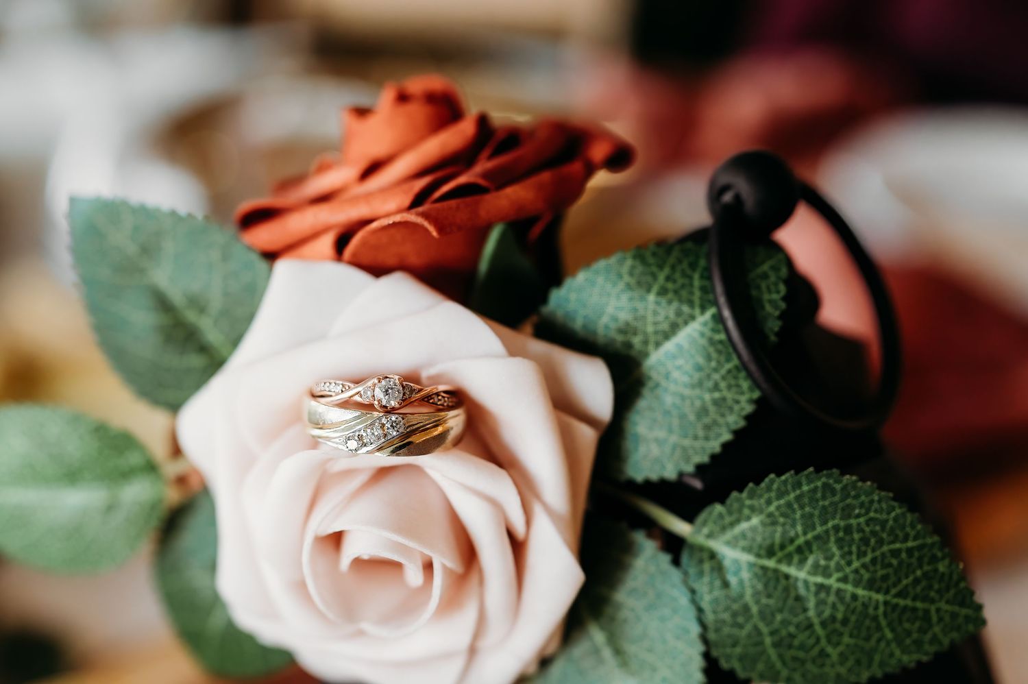 Close-up view of a delicate rose gold wedding ring resting on top of a soft pink rose with green leaves in background.