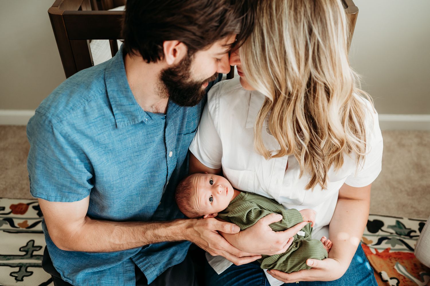 Parents share intimate moment while holding newborn baby in olive colored onesie at home.
