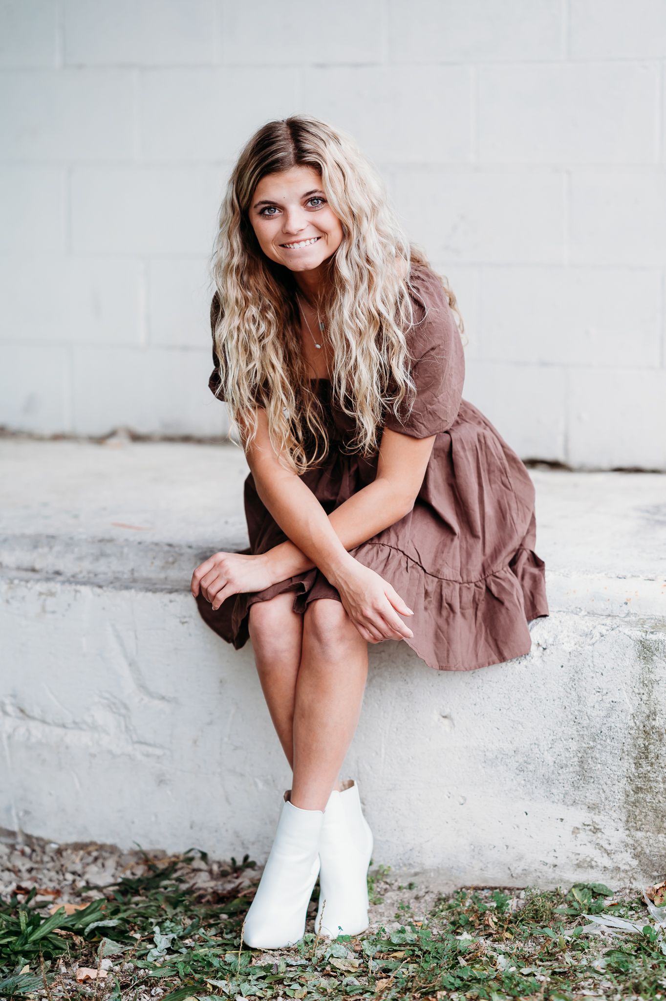 Person in a brown ruffled dress and white boots sits casually on a concrete ledge against a white wall.