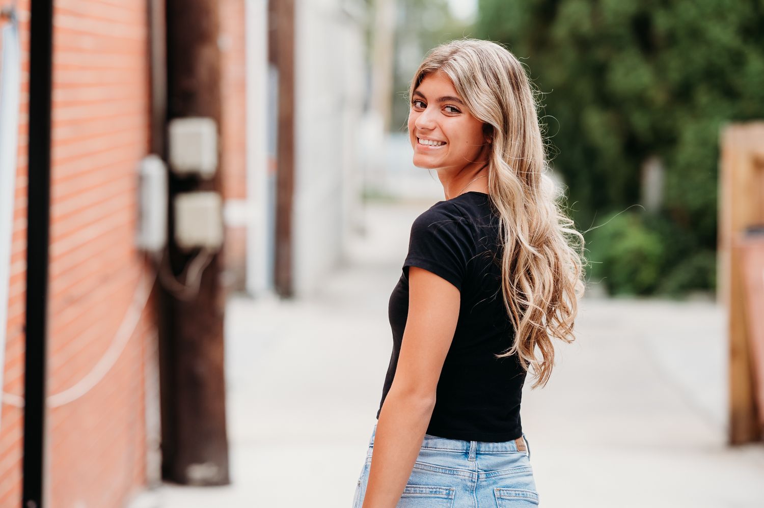 Woman with long blonde wavy hair wearing a black t-shirt and jeans looks over her shoulder while walking down a sidewalk.