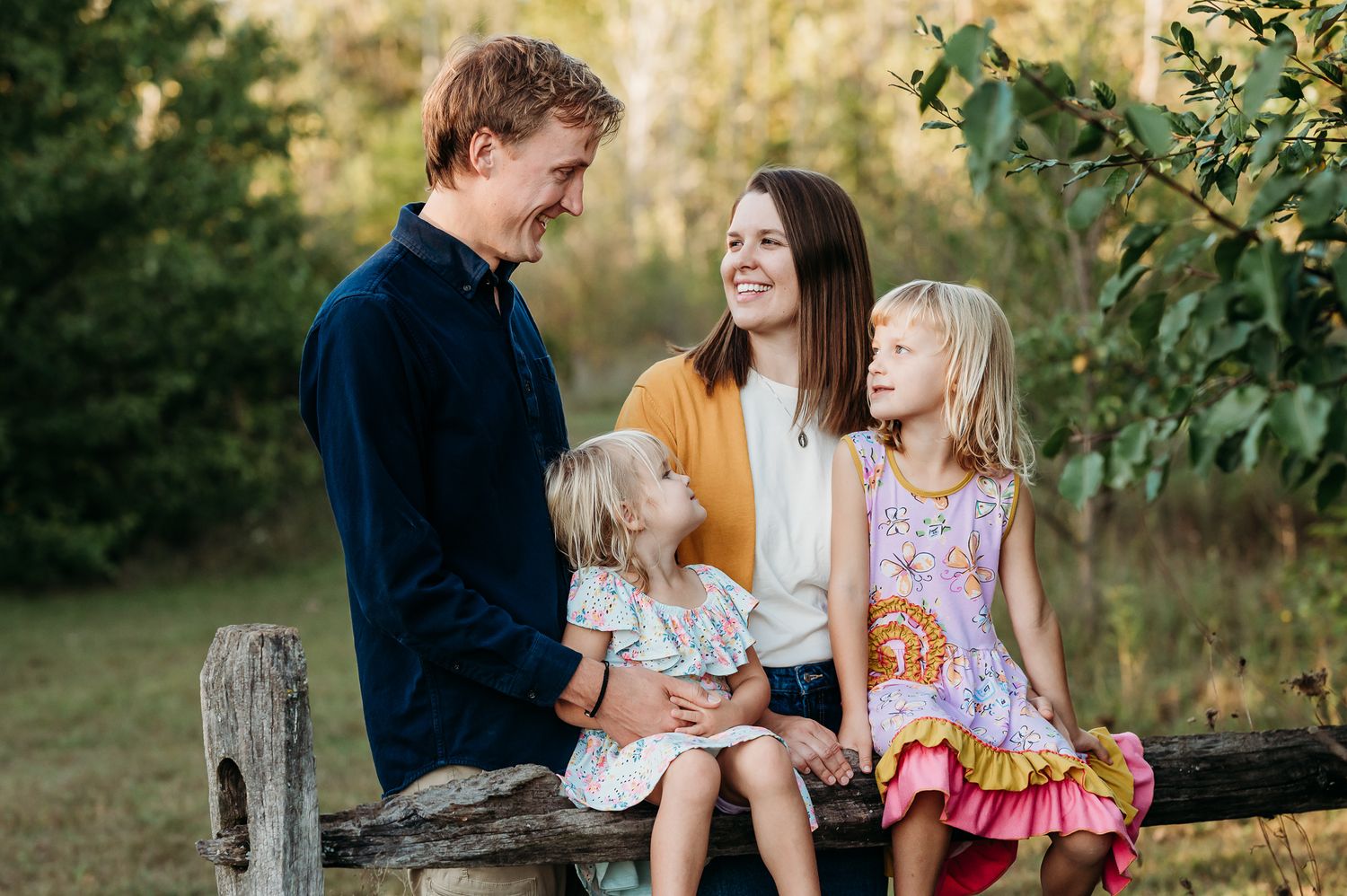 A family sits together outdoors on wooden fence during golden hour photo session at Washington Township Park in Avon, Indiana.