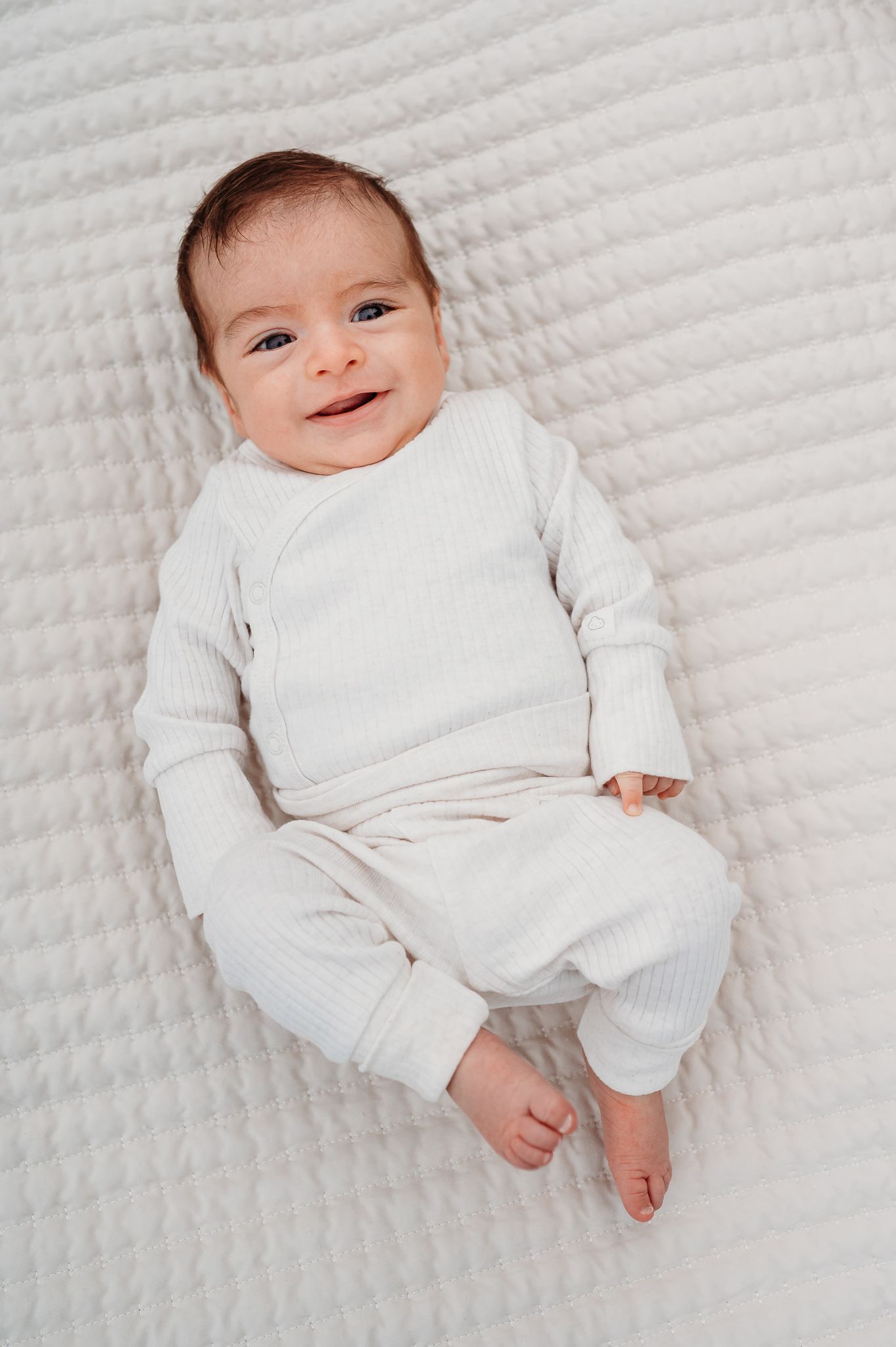 A baby in white clothing lies on a textured white blanket while looking at the camera with a sweet expression during outdoor newborn photos.