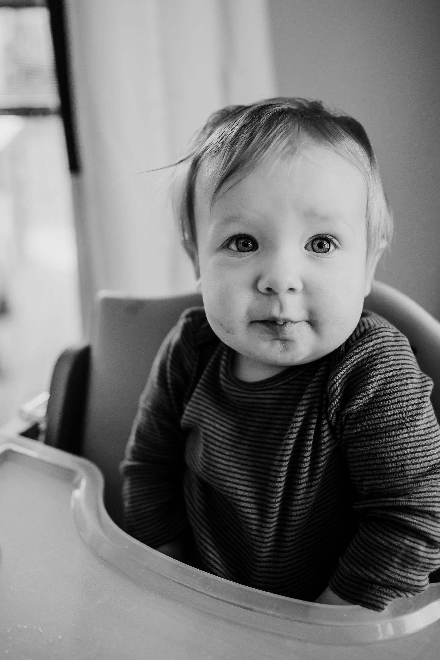 Black and white portrait of an adorable baby sitting in a high chair, wearing a striped shirt, making a cute face.
