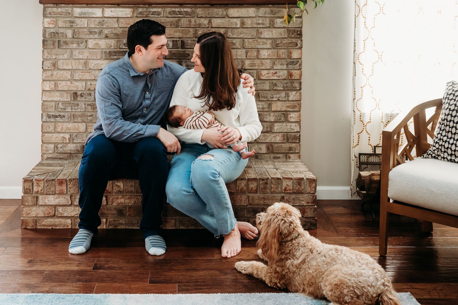 A couple sits on brick fireplace in their Noblesville home with their baby and a goldendoodle dog laying at their feet on hardwood floors.