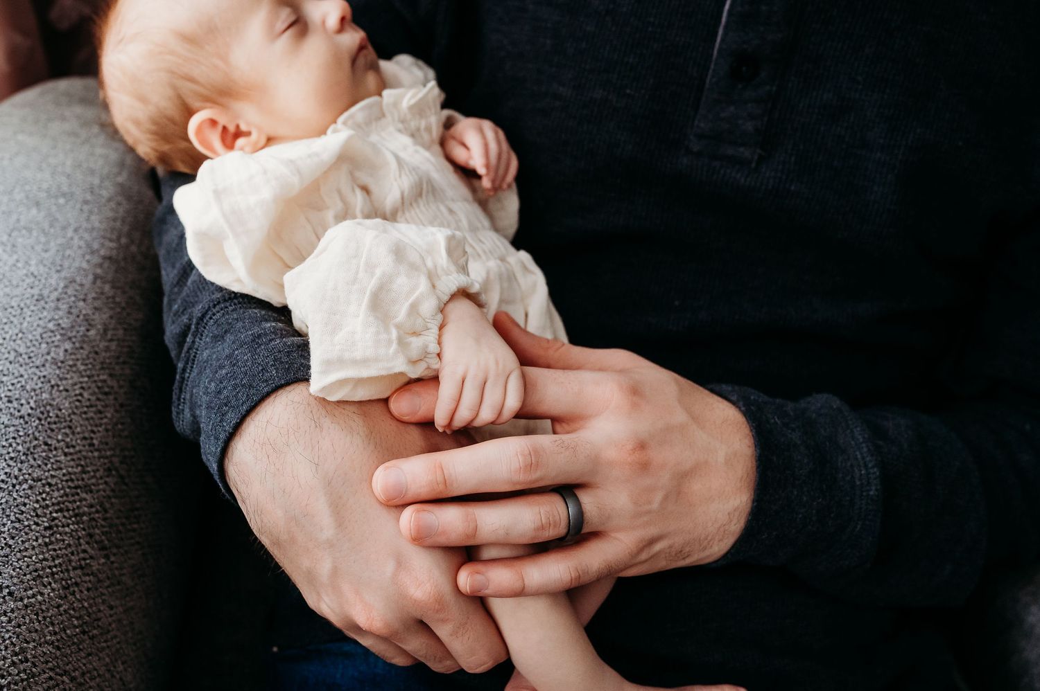 A baby in white clothing lies back while holding dad's finger.