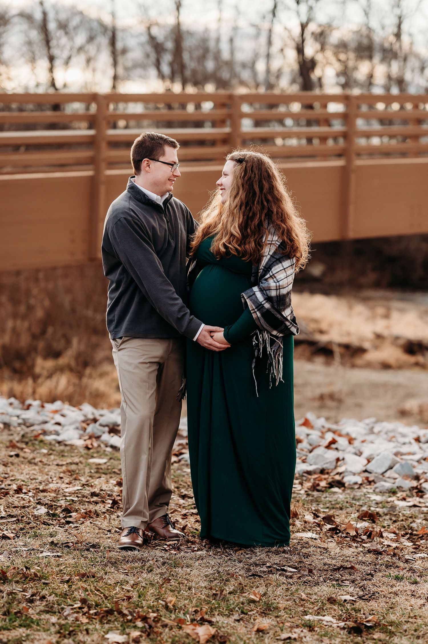 Pregnant couple holding hands outdoors in Plainfield, woman in green maternity dress with plaid shawl, wooden bridge background.