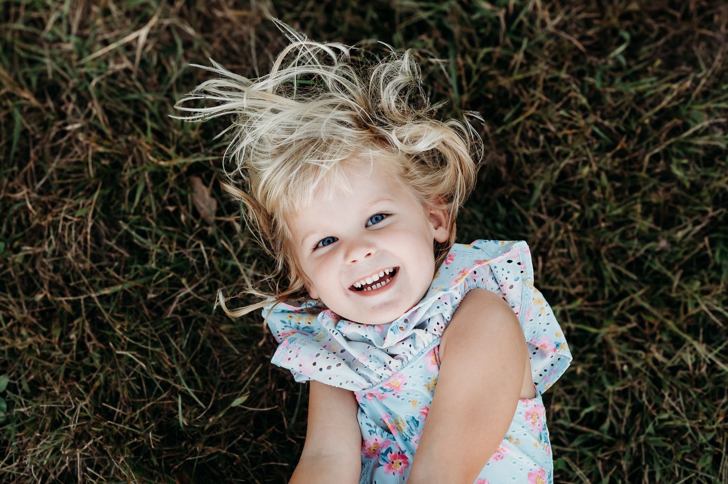 A child in a floral dress plays and laughs while sitting in grass outdoors during petite family photo session in Avon, Indiana.