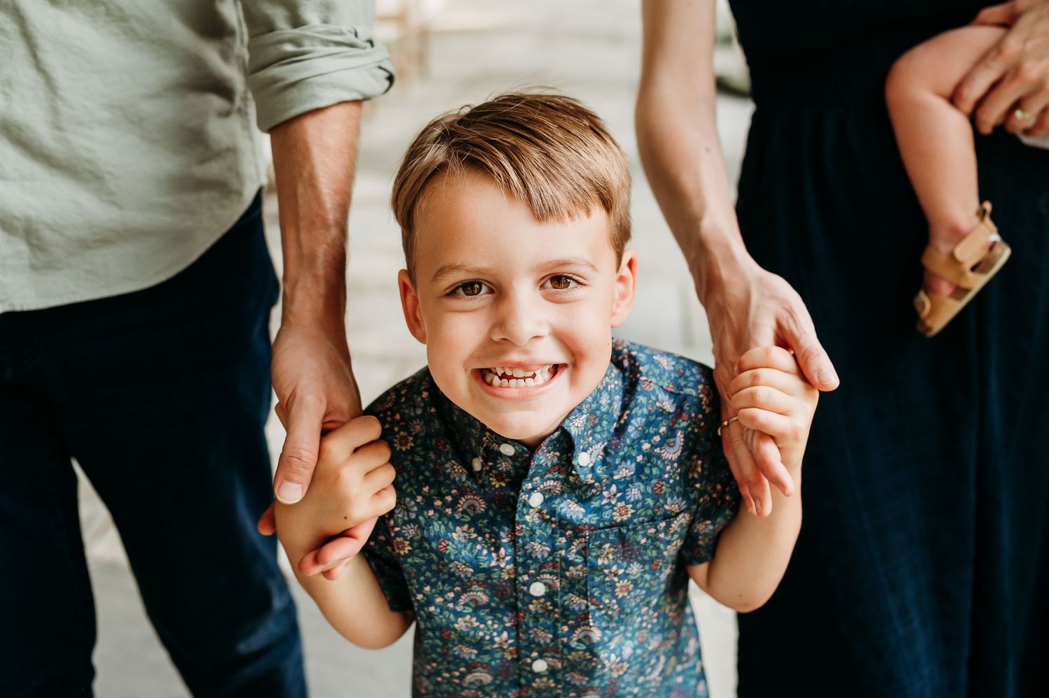 A young boy in a patterned shirt being playful while holding mom and dad's hands.