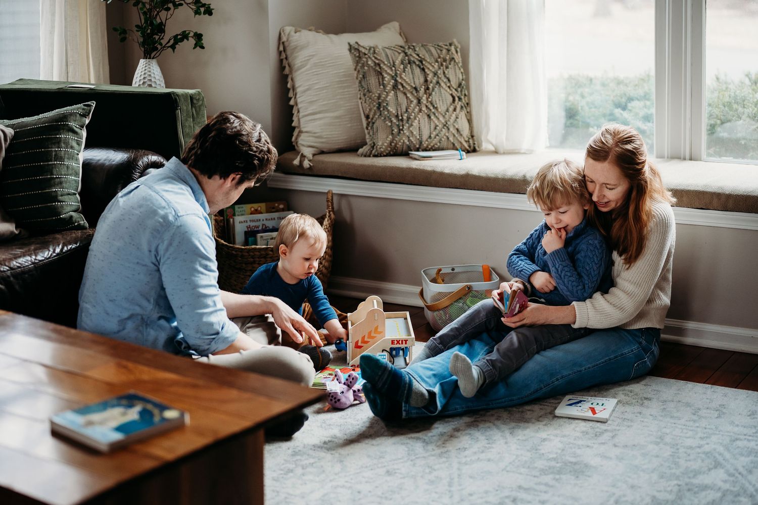 Family of four sitting on living room floor playing together near a coffee table and window seat.